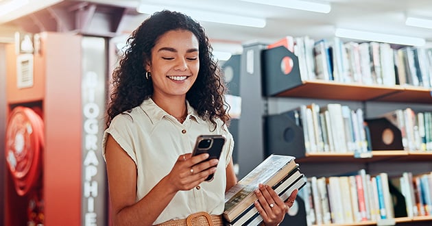 A young woman looks at her phone, holding a pile of books.