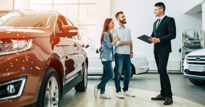 Car dealership showroom with an orange SUV in the foreground and a salesman talking to a couple.