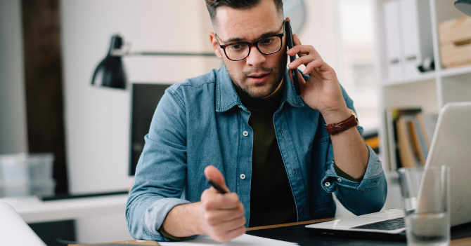 Man in glasses on a phone call at a desk, holding a pen near a laptop.