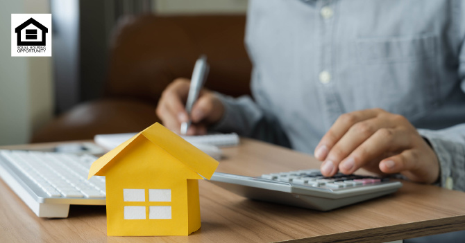 A small paper house on a table with a person using a calculator and pen in the background.