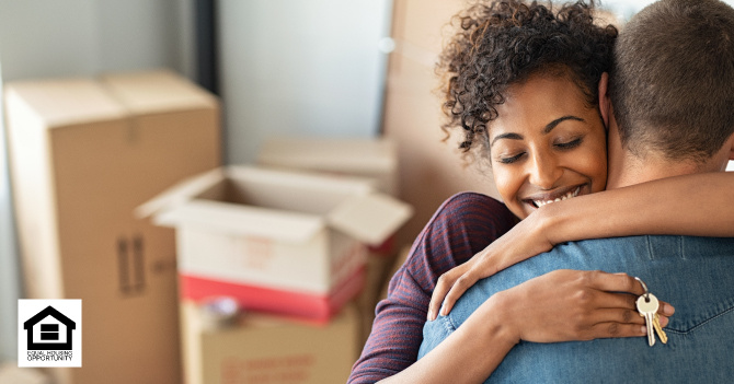 A smiling woman embraces a man holding a set of keys, with moving boxes in the background.