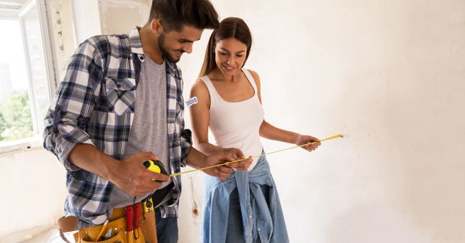 Man and woman measuring a wall with a yellow measuring tape in an unfinished room.