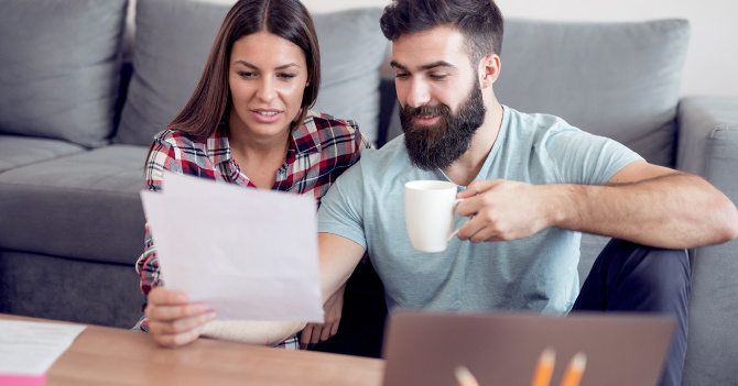 A woman and man seated on the floor, reviewing a piece of paper. The man holds a white mug.