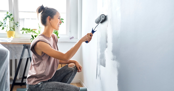 Woman kneeling and painting an interior wall with a roller near a window.