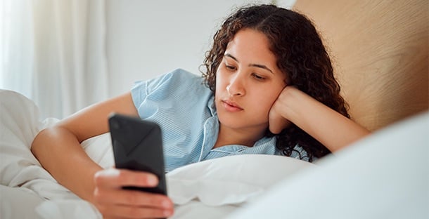 A woman is laying down on her bed looking contemplative at her smartphone.