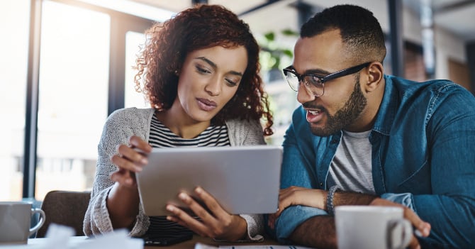 Two people looking at a tablet device in a bright setting with coffee mugs on the table.