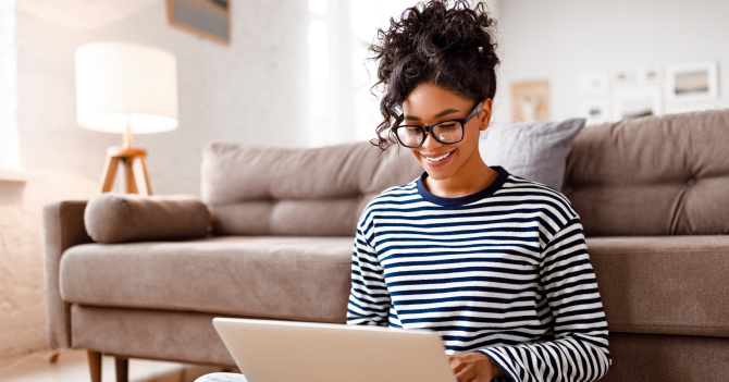 Person with curly hair smiling while using a laptop in a living room with a sofa and lamp.