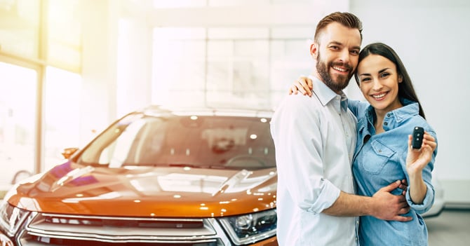 A smiling couple in a showroom stands beside a new orange car; the woman holds a car key fob.